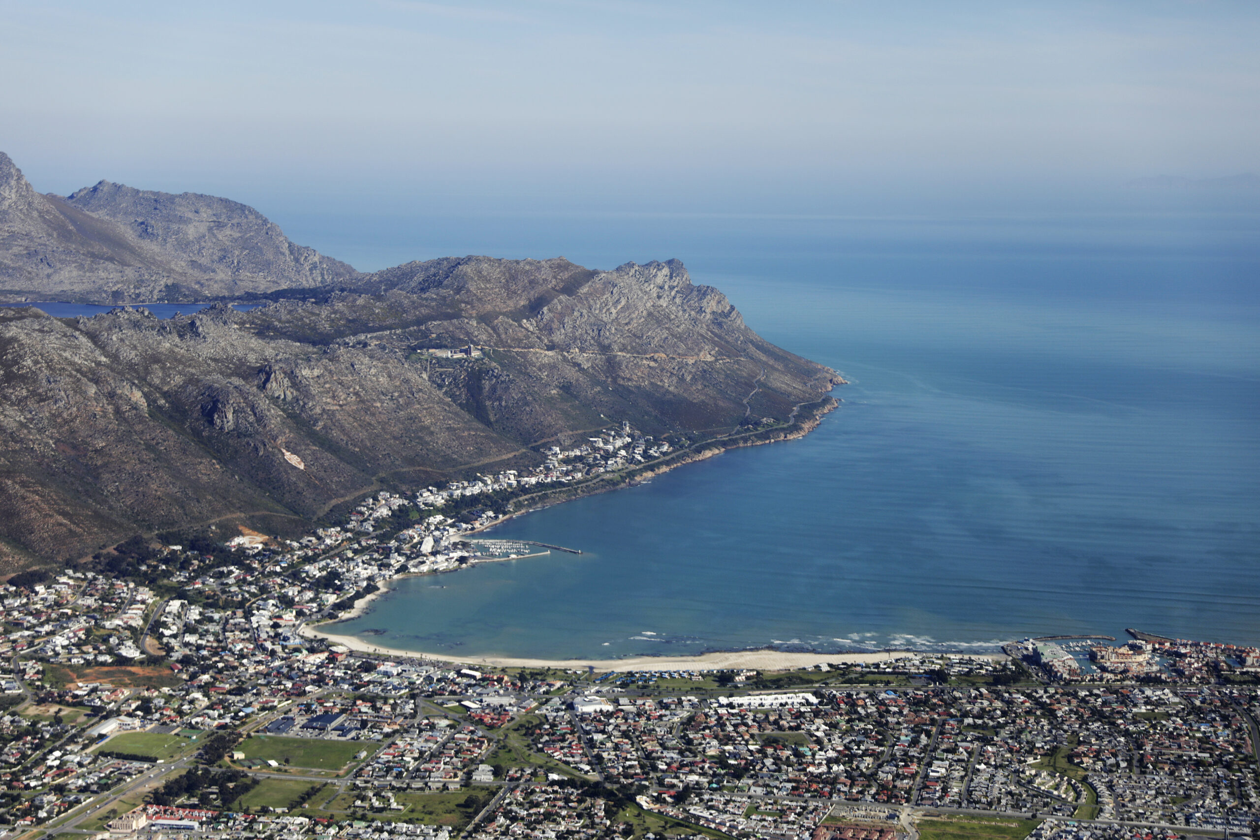 erial view of a coastal town at the foot of rugged mountains. A curved sandy beach and a small harbour sit on calm blue bay water.